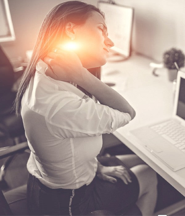 Woman sitting at a desk holding her neck in discomfort, illustrating neck strain and tension from poor posture while working on a computer.